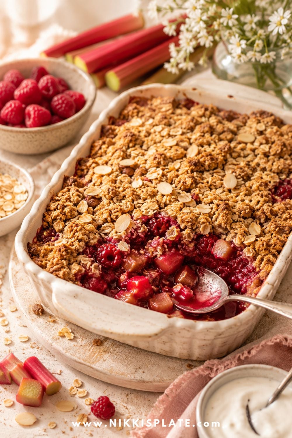 Healthy rhubarb raspberry crisp in a white baking dish with golden oat crumble, fresh raspberries, rhubarb stalks, and yogurt on the side.