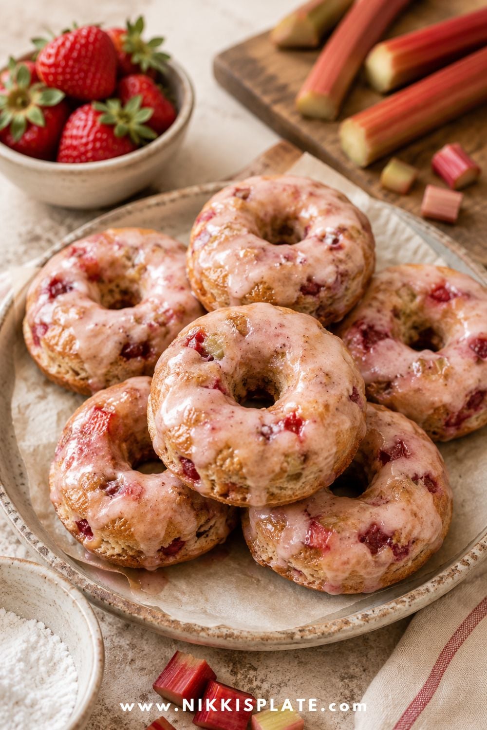 Healthy strawberry rhubarb baked donuts with pink glaze, fresh strawberries, rhubarb stalks, chopped rhubarb, and powdered sugar.