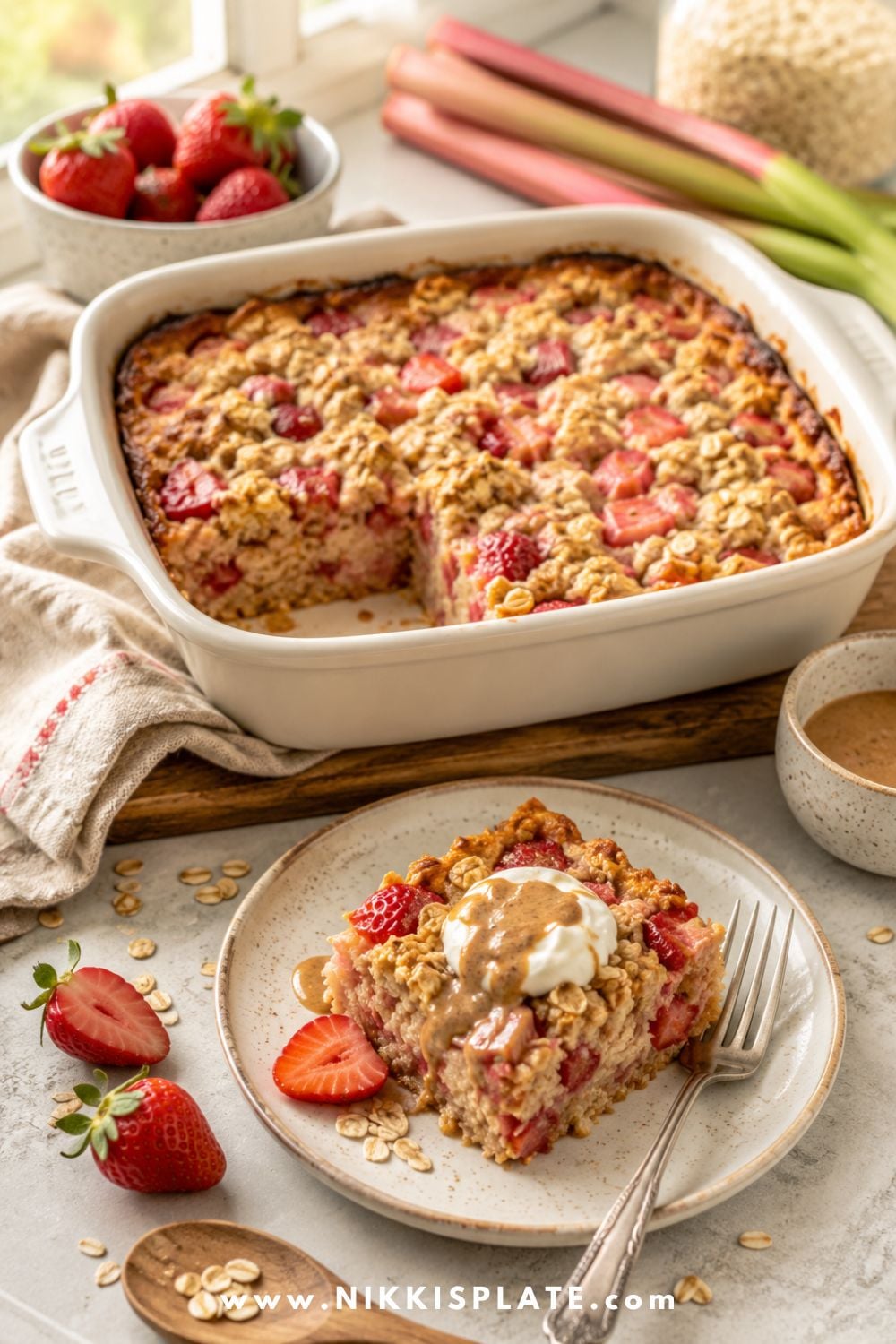 Healthy strawberry rhubarb baked oatmeal in a white baking dish with a plated slice topped with yogurt, almond butter, fresh strawberries, oats, and rhubarb.