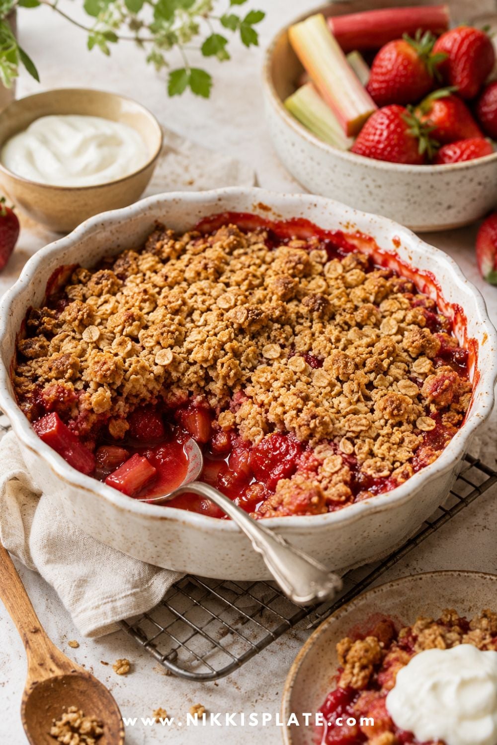 Healthy strawberry rhubarb crisp in a white baking dish with oat crumble topping, fresh strawberries, rhubarb stalks, and yogurt on the side.