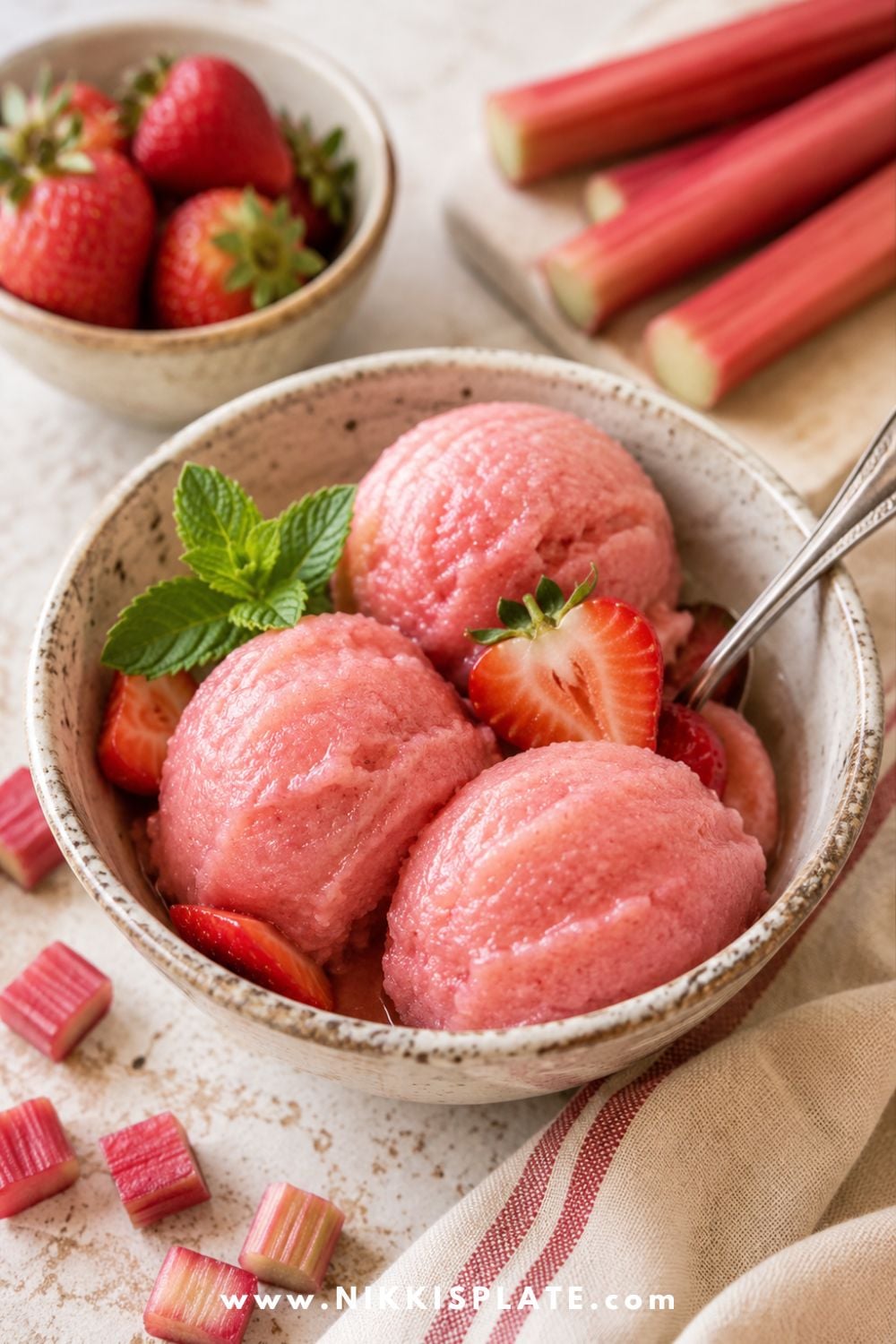Healthy strawberry rhubarb sorbet scoops in a ceramic bowl with fresh strawberries, mint, rhubarb stalks, and chopped rhubarb.