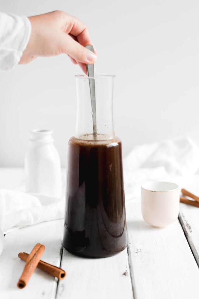 Large tumbler of overnight cinnamon cold brew coffee, hand stirring with spoon, almond milk in jar behind, cinnamon sticks beside