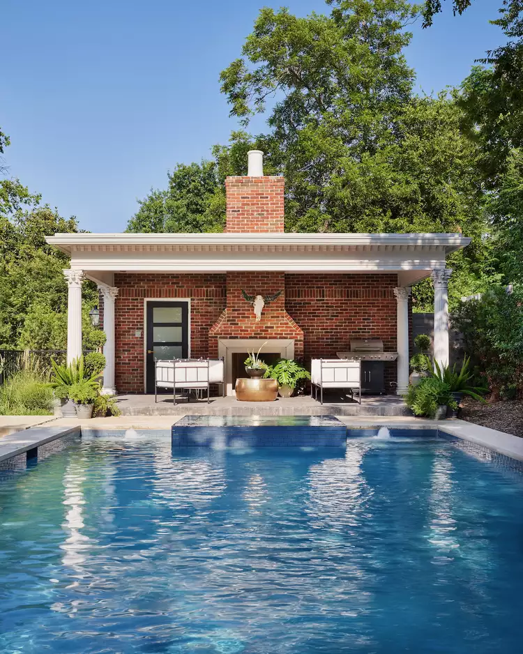 classic red brick pool house with white columns and an outdoor fireplace beside a rectangular backyard swimming pool