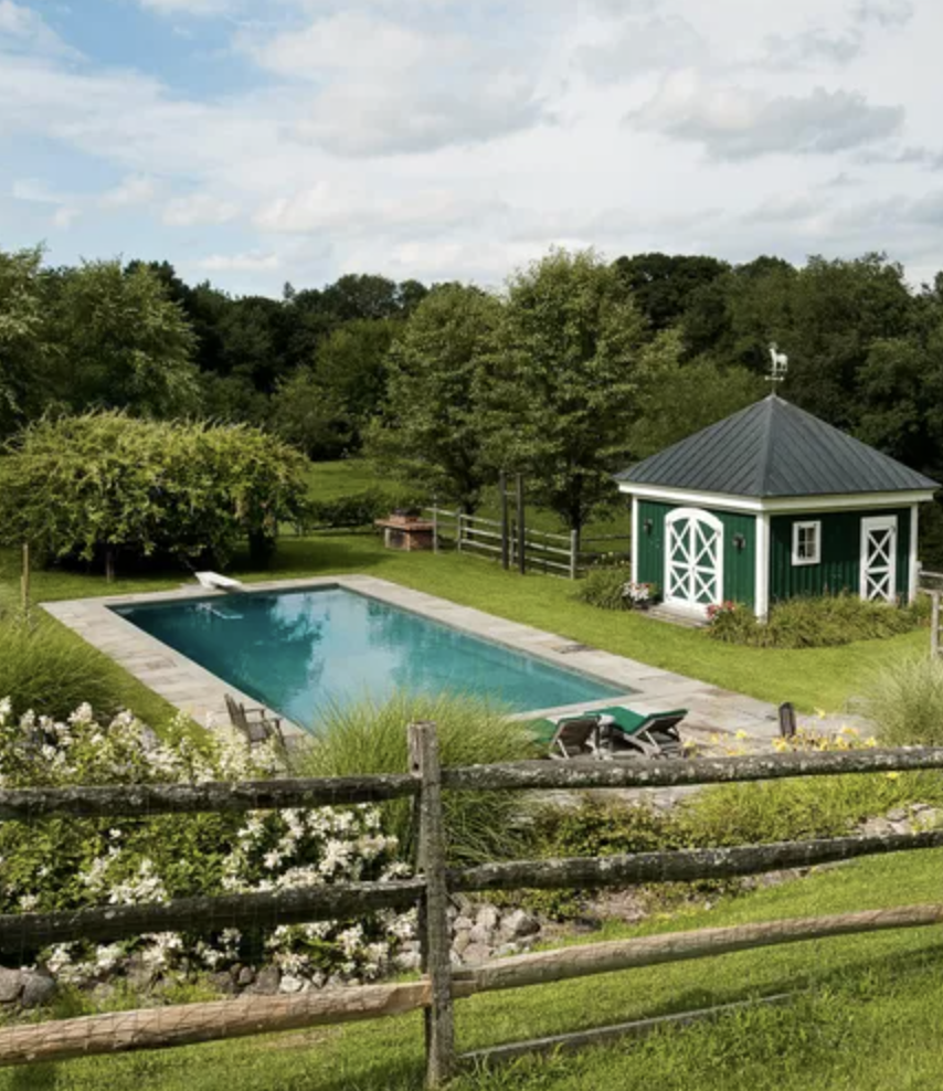 green barn-style pool house with white crossbuck doors beside a rectangular backyard pool surrounded by wooden fencing and lush countryside landscaping
