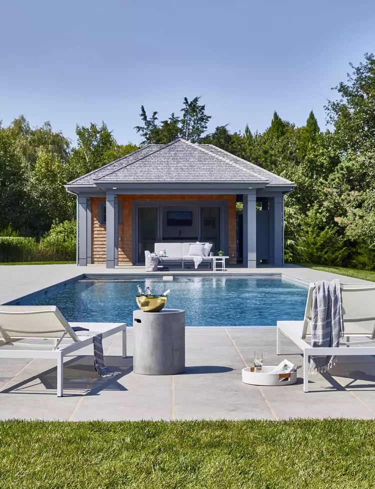 modern pool house with gray siding, cedar accents, hip roof, and a rectangular backyard swimming pool with white lounge chairs