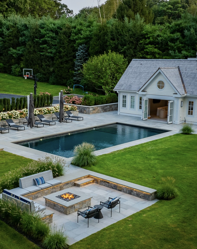 modern backyard with white pool house, rectangular swimming pool, lounge chairs, and sunken fire pit seating area