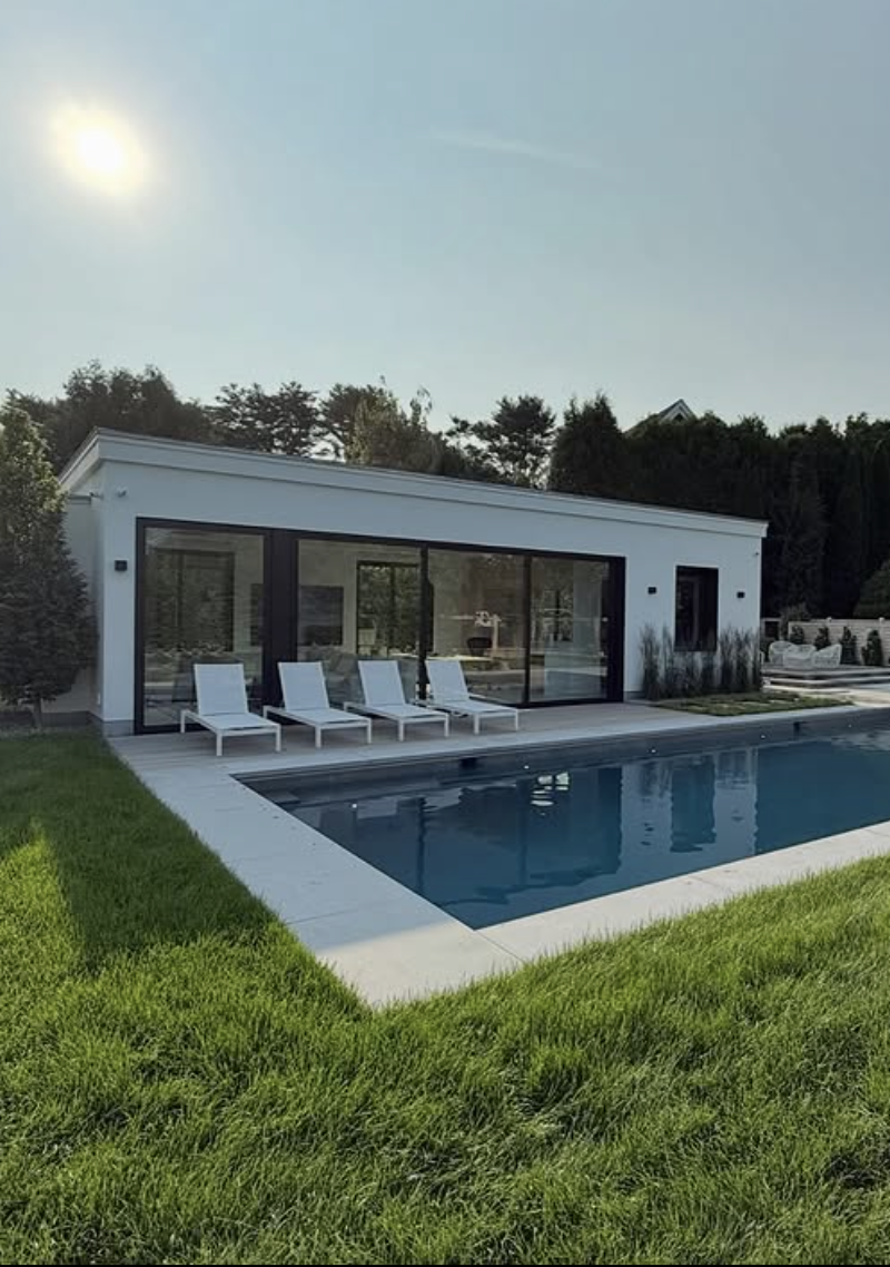 modern white flat roof pool house with black framed sliding glass doors and minimalist lounge chairs beside a rectangular backyard pool