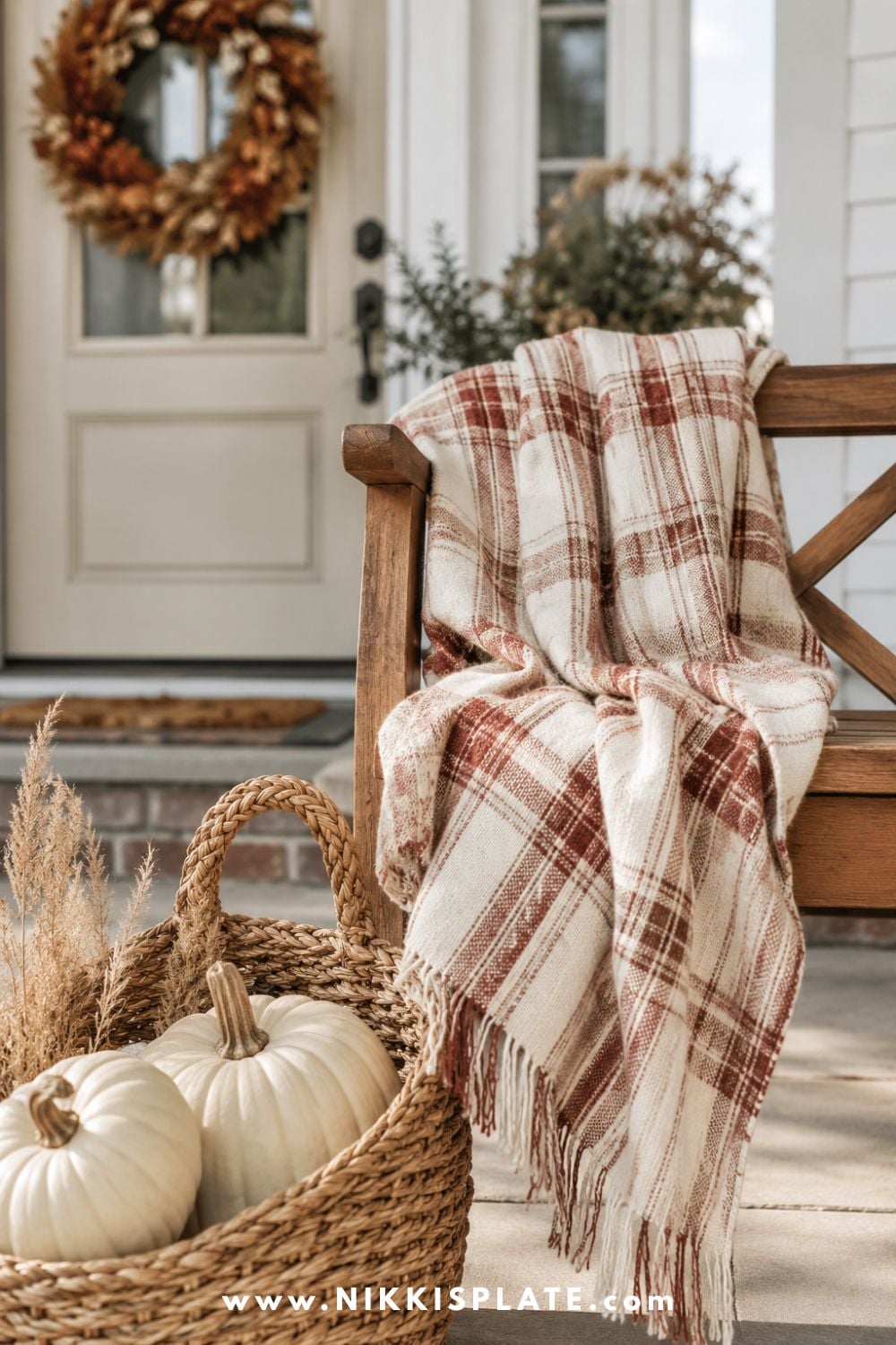 Cream and rust plaid throw draped over a wooden porch bench beside a basket of white pumpkins.