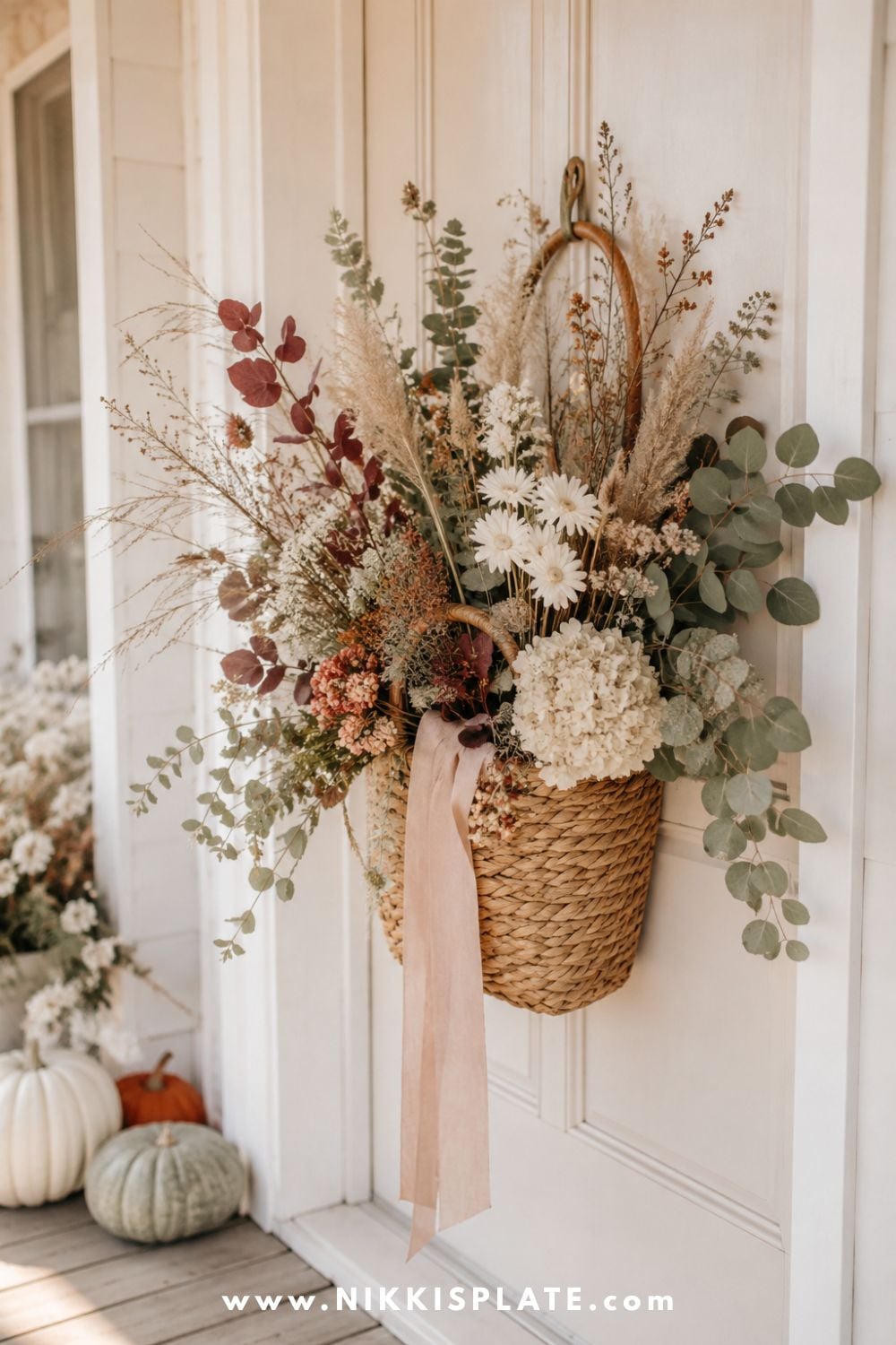 Woven fall door basket filled with eucalyptus, dried grasses, burgundy leaves, cream flowers and muted orange stems.
