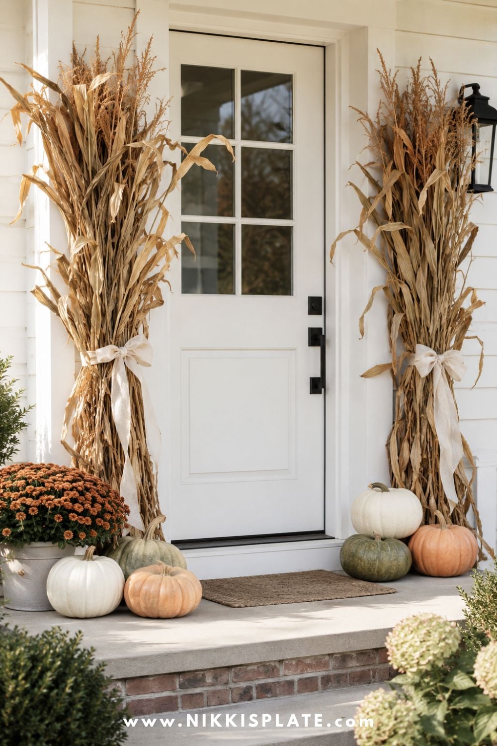 White farmhouse front door framed with cornstalk bundles, cream pumpkins and rust-colored mums.