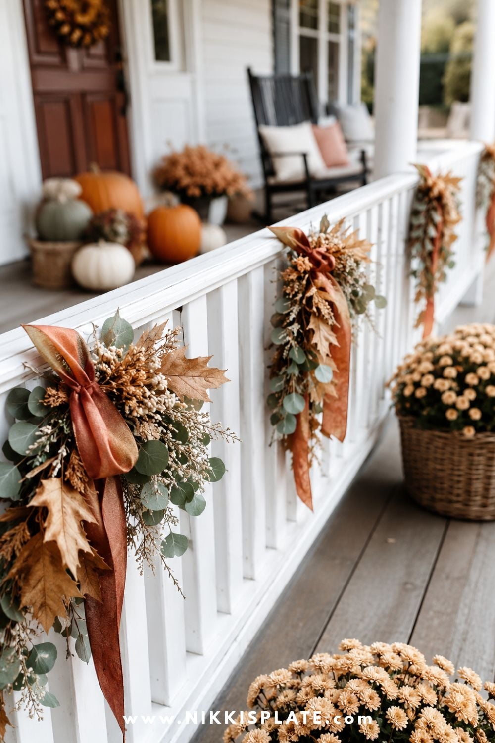 White porch railing decorated with fall leaf swags, muted rust ribbon and small baskets of mums.