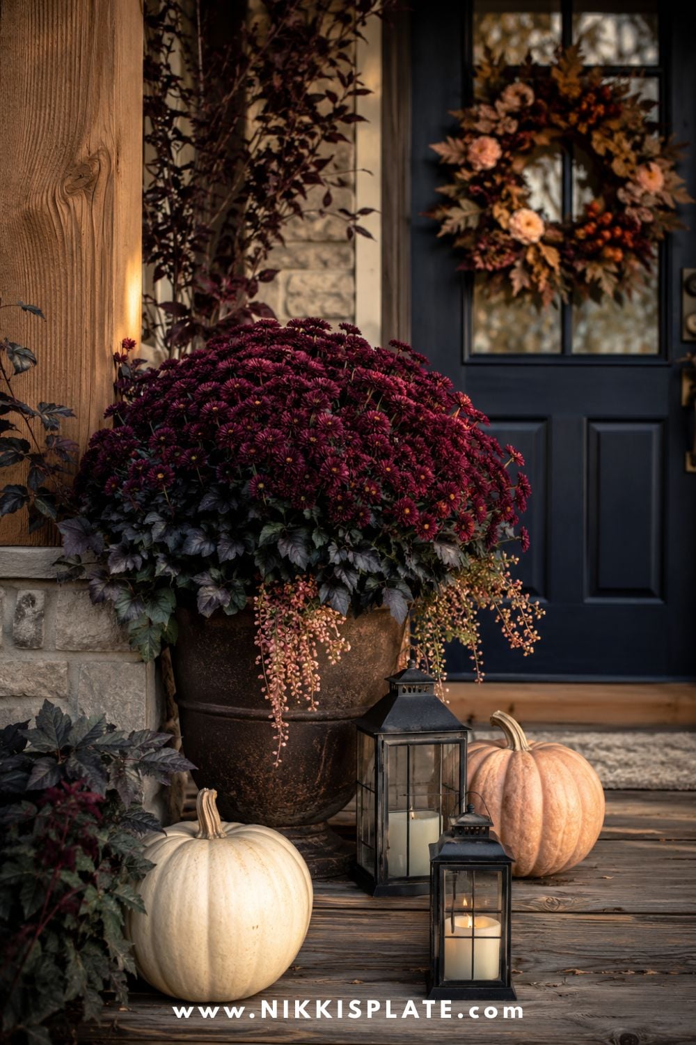 Moody fall porch styled with burgundy mums, cream pumpkins, black lanterns and a navy front door.