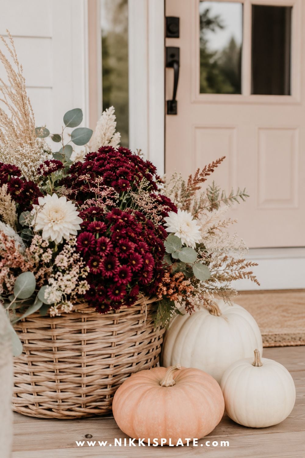 Large woven basket filled with burgundy mums, cream flowers, eucalyptus and dried grasses beside a front door.