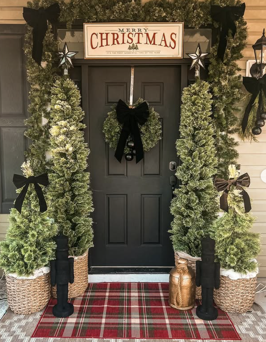 Christmas porch with black door, greenery garland, black ribbon wreath, plaid rug, tall potted trees, and nutcracker statues.