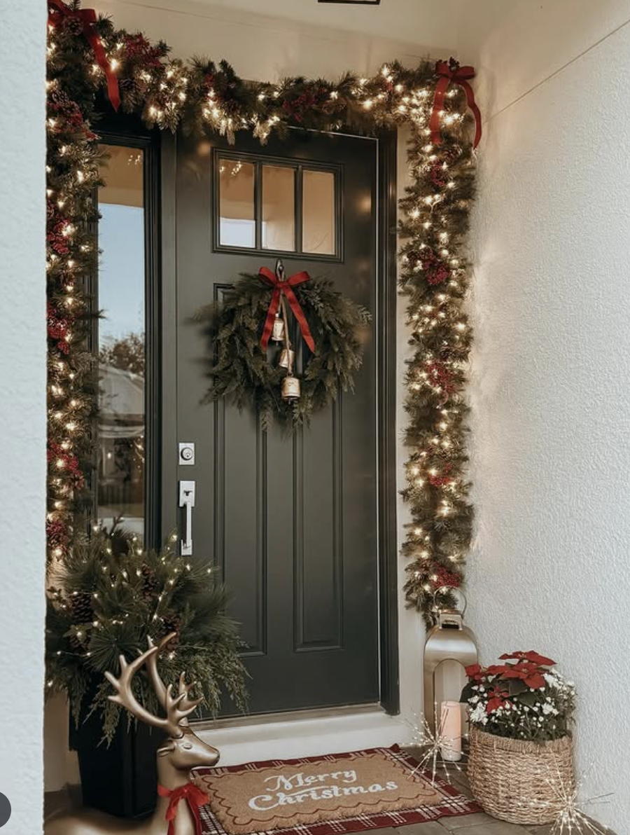Christmas front porch with black door, lit garland, red bows, greenery wreath with bells, reindeer figurine, and poinsettia plant.