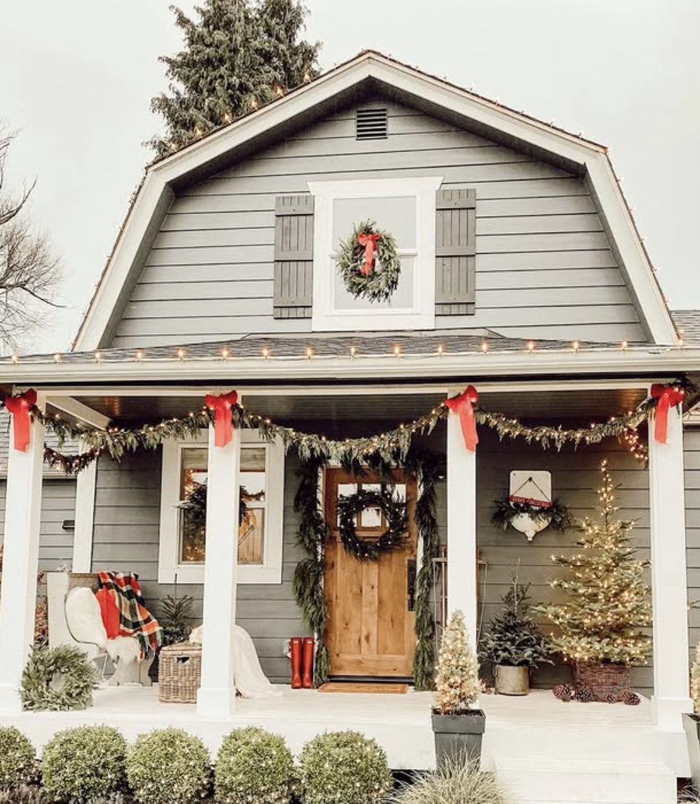 Farmhouse front porch with greenery garland and red bows, wooden front door with wreath, plaid blanket on chair, and lit Christmas tree.