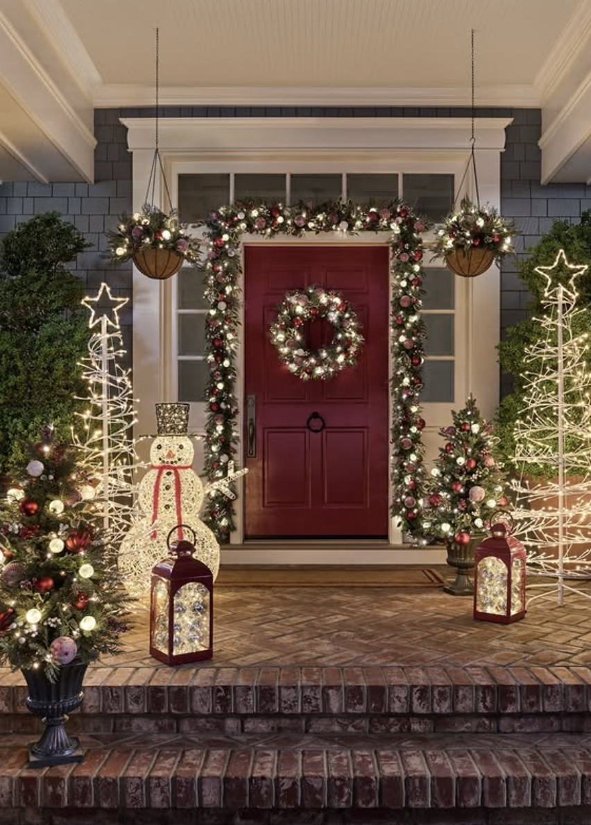 Front porch with red door, garland and wreath with lights, glowing snowman, star-shaped light trees, mini decorated trees, and lanterns.