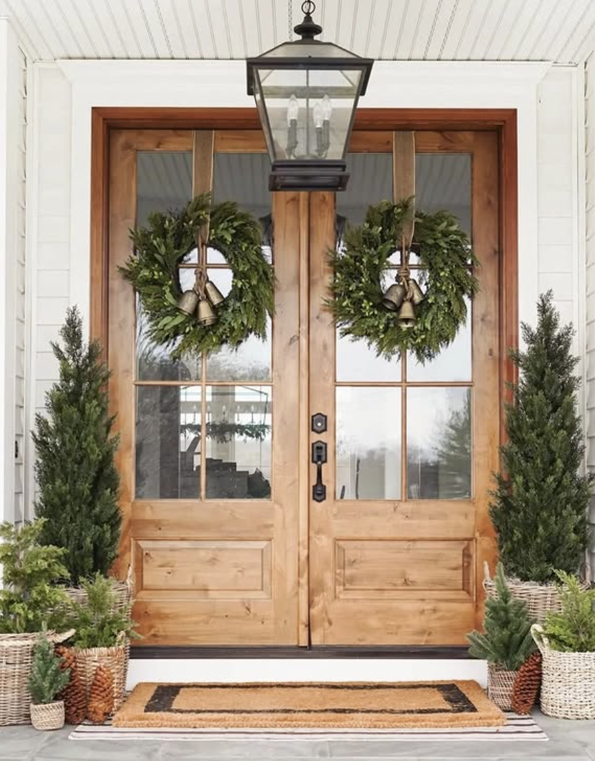 Front porch with double wooden doors, simple greenery wreaths with brass bells, tall potted evergreens, and woven baskets.