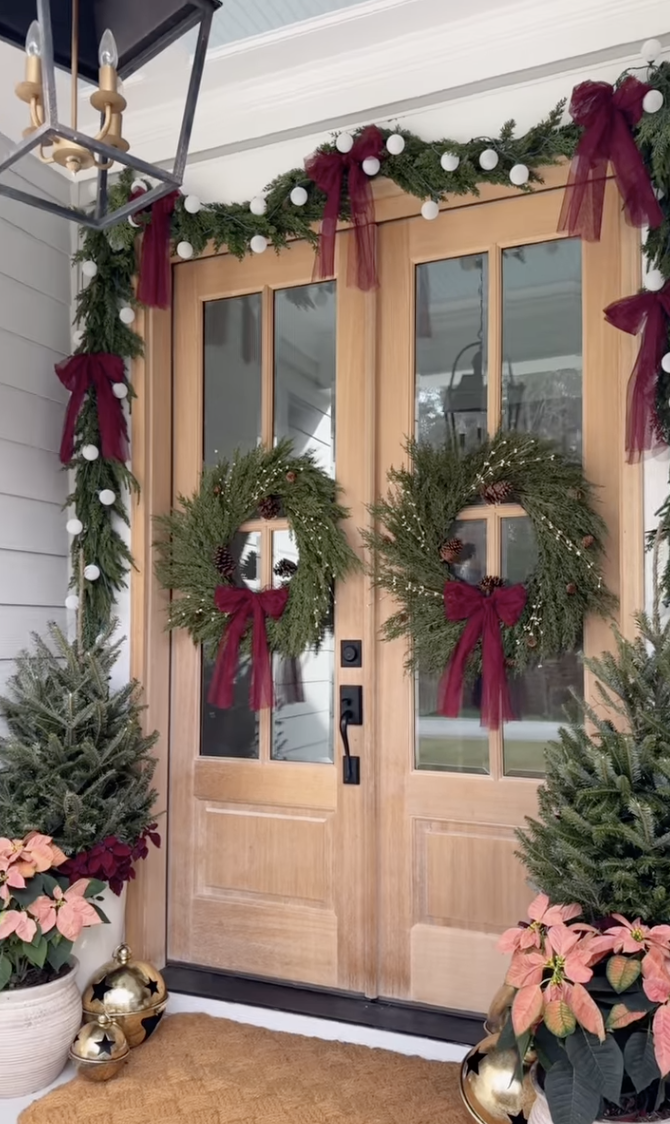 Front porch with double wooden doors, fresh garland with burgundy bows, matching wreaths, potted evergreens, and poinsettias.