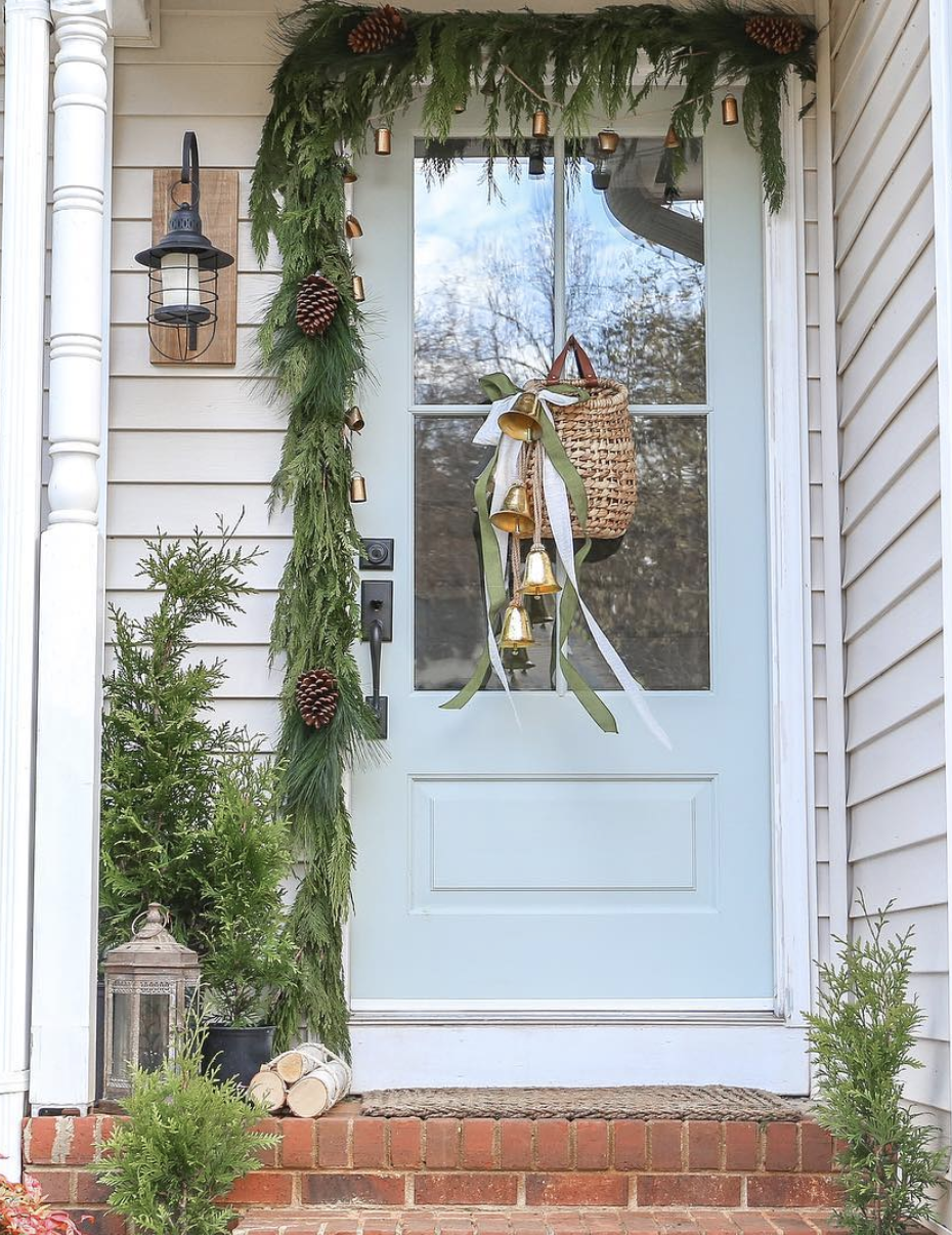 Light blue front door with natural pine garland, pinecones, woven basket with gold bells, and small potted evergreens.