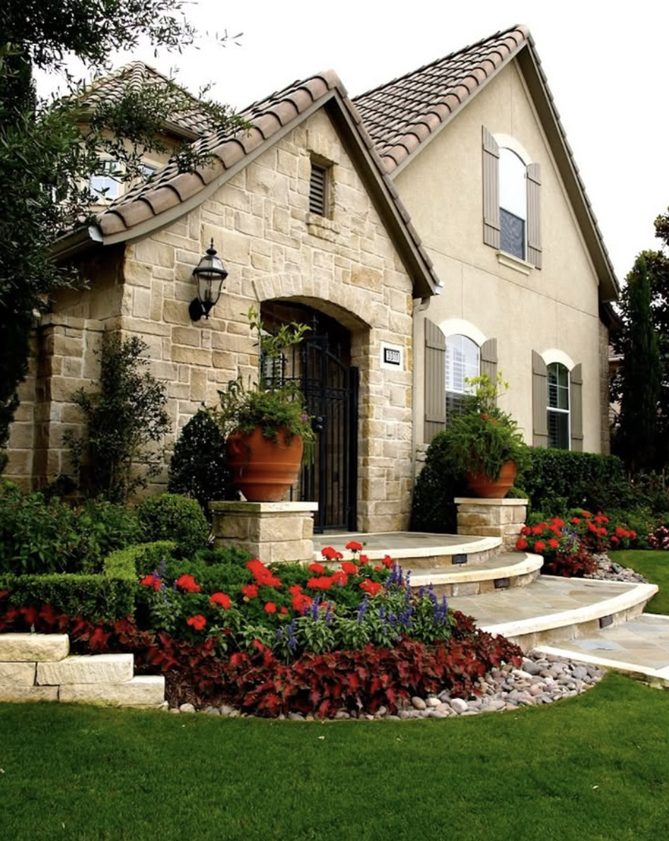 Landscaping ideas for front of house featuring stone steps, terracotta planters, red flowering plants, and manicured shrubs.