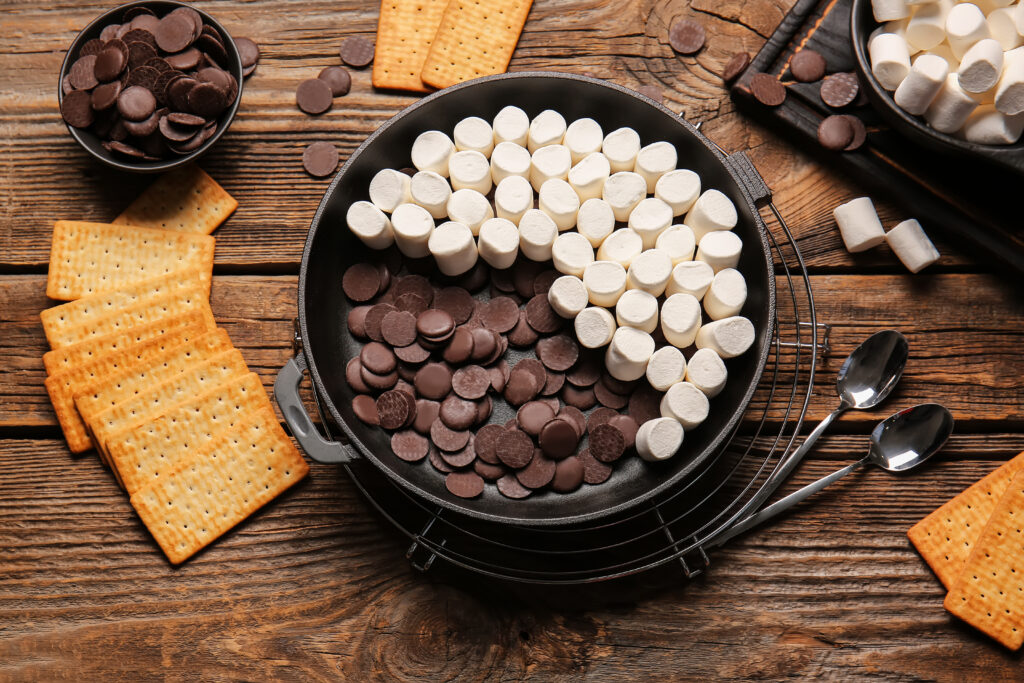 Frying pan with uncooked S'mores dip on wooden background- Easy oven baked s’mores dip recipe; This easy oven baked s’mores dip recipe is perfect for summer BBQs, potlucks and parties. Made with marshmallows, chocolate chips and graham crackers! Crowd pleasing dessert dip!