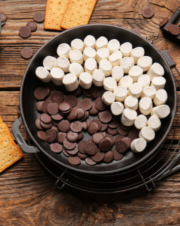 Frying pan with uncooked S'mores dip on wooden background- Easy oven baked s’mores dip recipe; This easy oven baked s’mores dip recipe is perfect for summer BBQs, potlucks and parties. Made with marshmallows, chocolate chips and graham crackers! Crowd pleasing dessert dip!