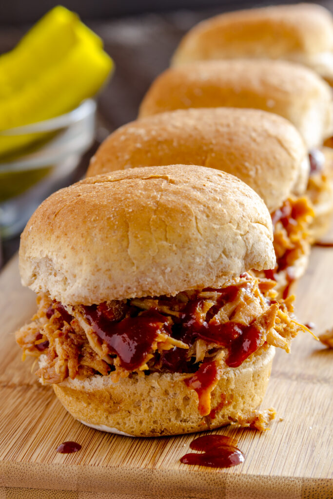 Row of pulled pork barbeque sliders sitting on wooden cutting board with red striped napkin and dill pickles in background - 10 Easy Campfire Recipes (Be the Master of Campfire Cooking); Here are 10 campfire cooking recipes to try this summer!