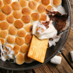 Frying pan with tasty S'mores dip and crackers on wooden background - Easy oven baked s’mores dip recipe; This easy oven baked s’mores dip recipe is perfect for summer BBQs, potlucks and parties. Made with marshmallows, chocolate chips and graham crackers! Crowd pleasing dessert dip!