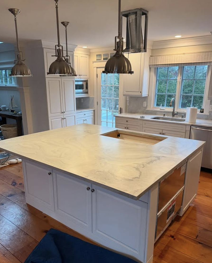White kitchen with large center island featuring a light-colored concrete countertop and industrial pendant lighting.