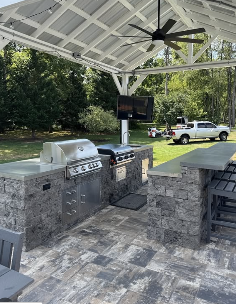 Covered outdoor kitchen with concrete countertops, stone base, built-in grill, bar seating, and ceiling fan.
