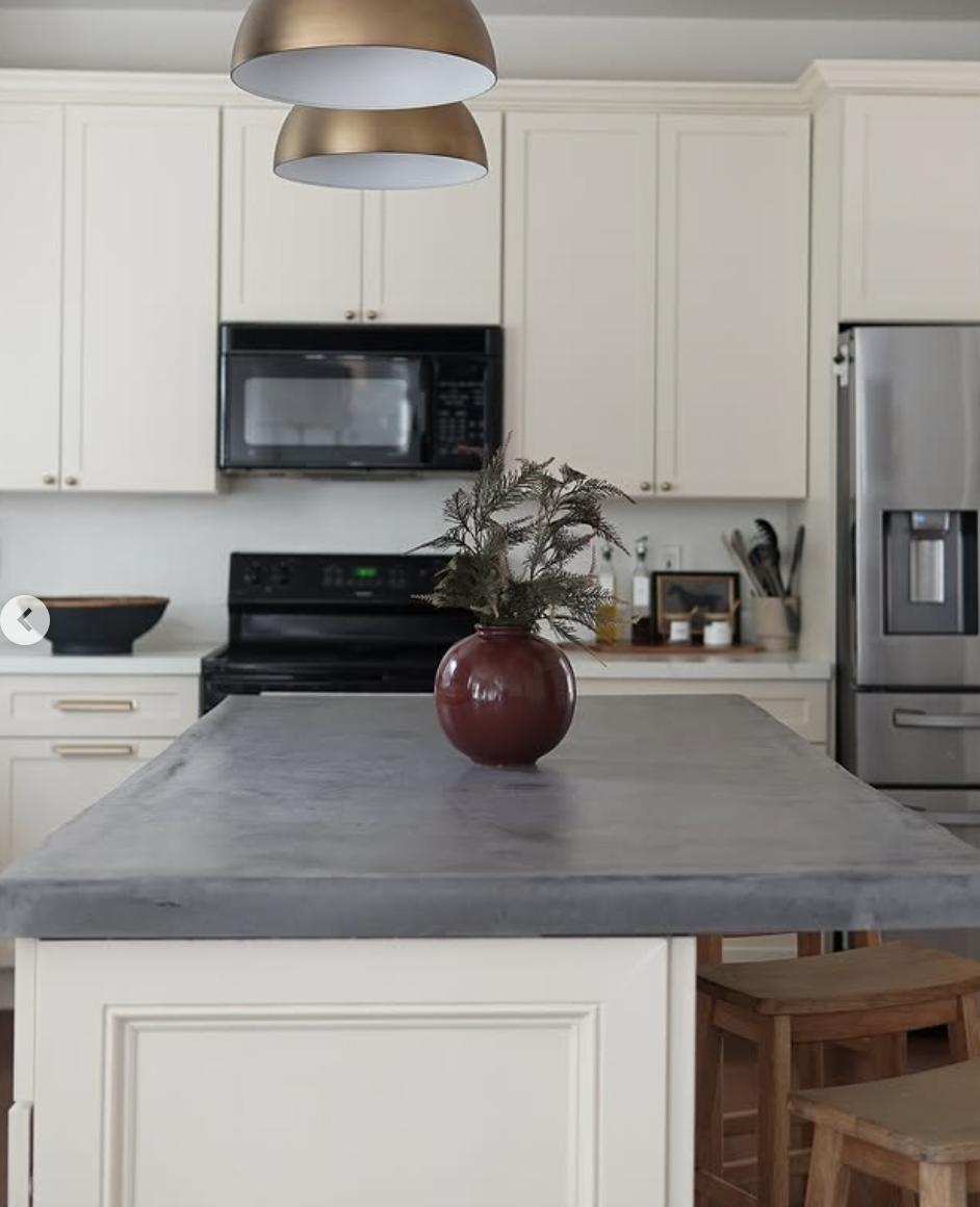 Minimalist cream kitchen with a smooth concrete island countertop, black appliances, and brass pendant lights above.