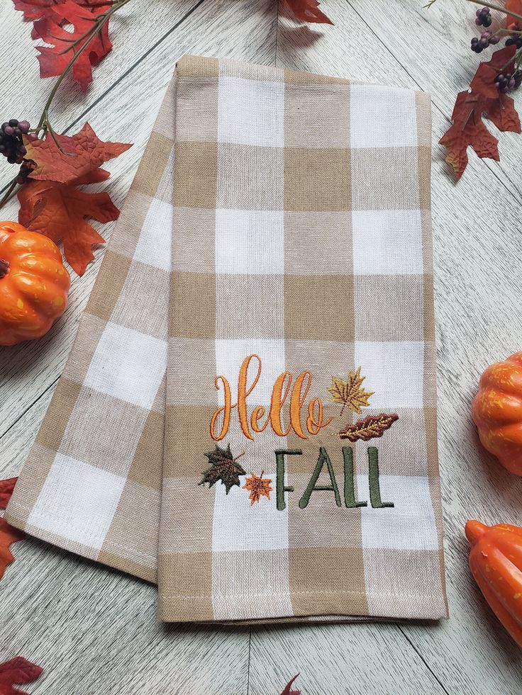 Budget-friendly fall kitchen update with a tan and white gingham dish towel embroidered with Hello Fall, surrounded by pumpkins and autumn leaves.