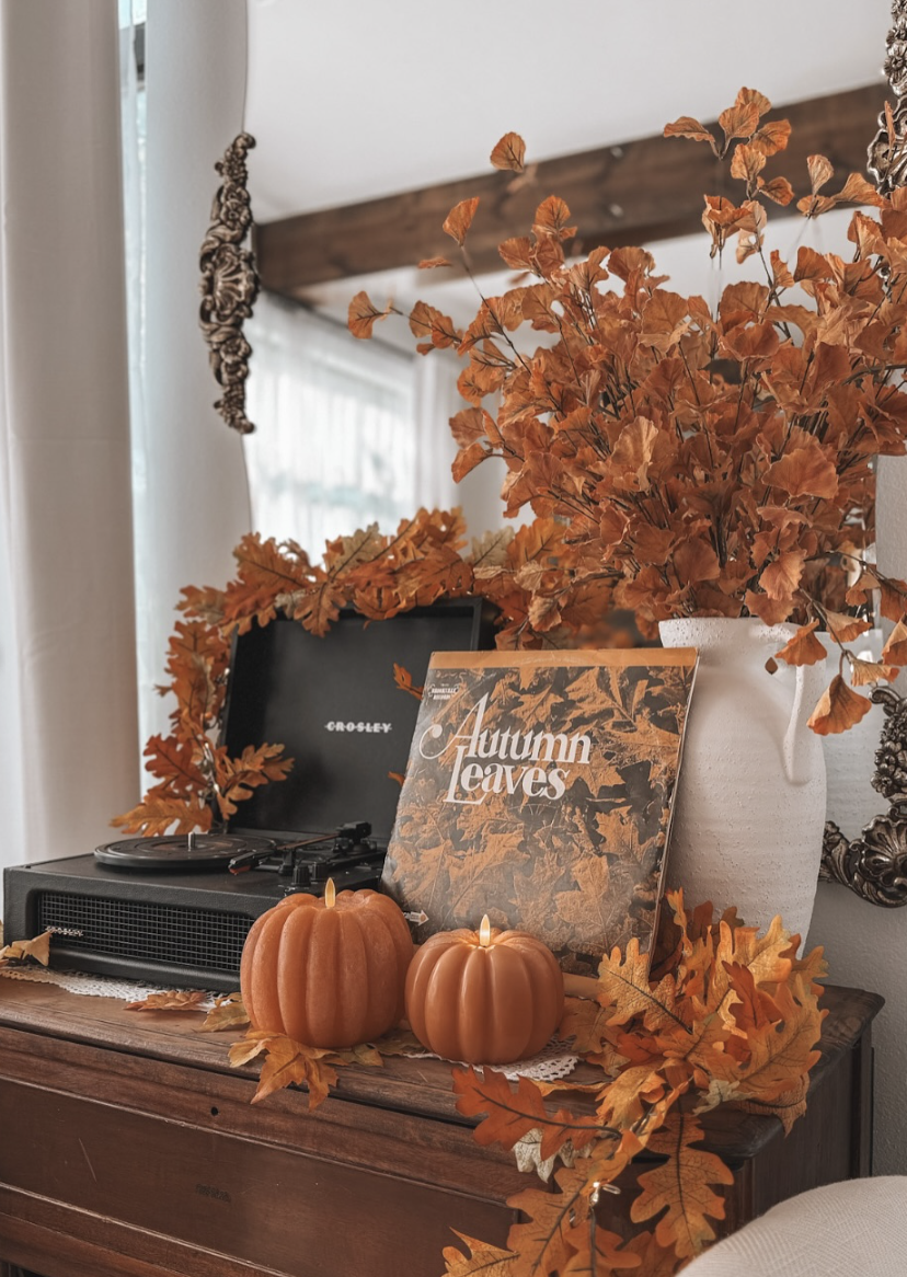 Cozy fall vignette with vinyl record player, autumn leaves in a white vase, mini pumpkins, and warm seasonal accents on a wooden console table.