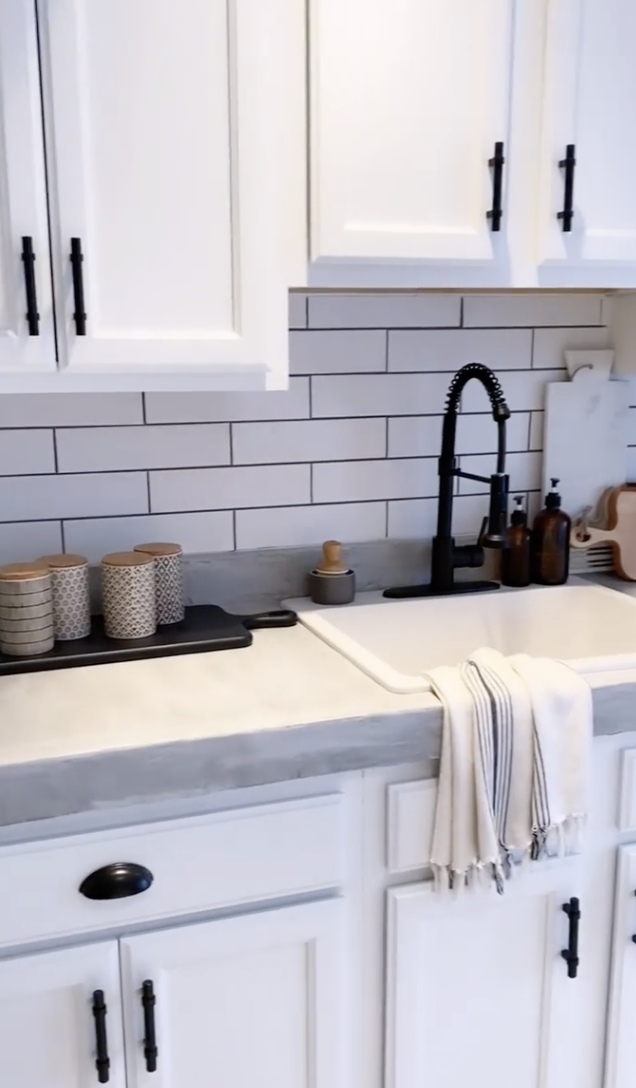 White farmhouse kitchen with shaker cabinets, subway tile backsplash, matte black faucet, and concrete countertops with a soft gray finish.