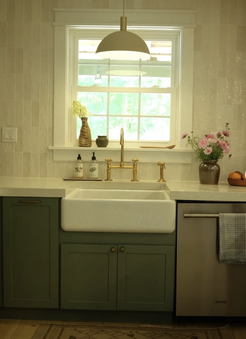 Farmhouse kitchen sink with brass faucet, muted green lower cabinets, creamy backsplash tile, and fresh flowers on the counter.