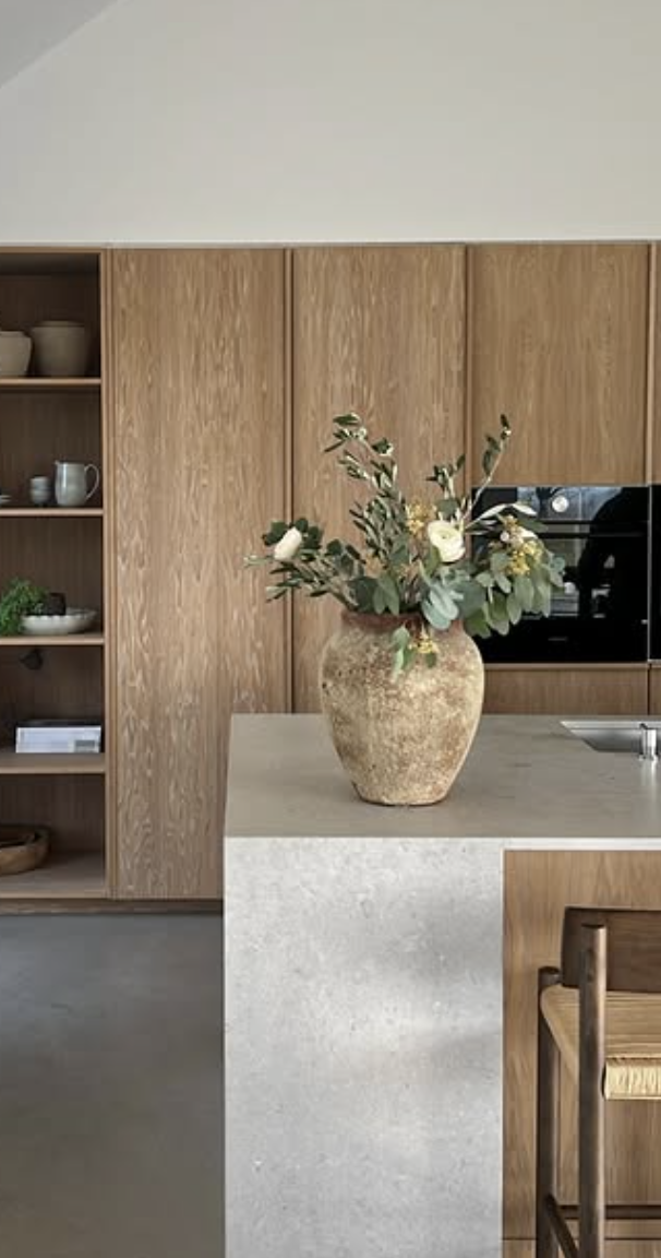 Minimalist kitchen with light wood cabinets, smooth concrete island countertop, and rustic ceramic vase centerpiece.