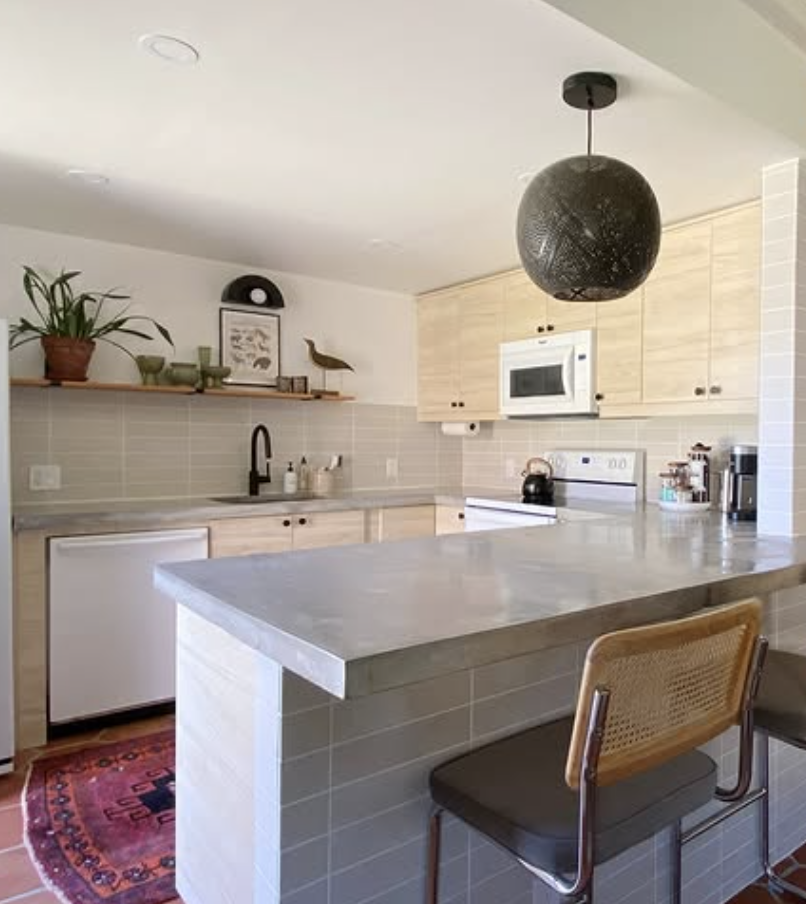 Minimalist kitchen with light wood cabinets, gray concrete countertops, open shelving, and a modern globe pendant light.