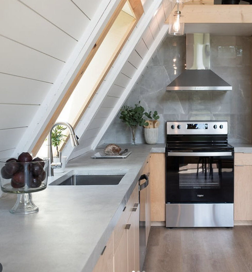 Bright A-frame kitchen with sleek concrete countertops, light wood cabinetry, stainless steel appliances, and a minimalist gray backsplash.