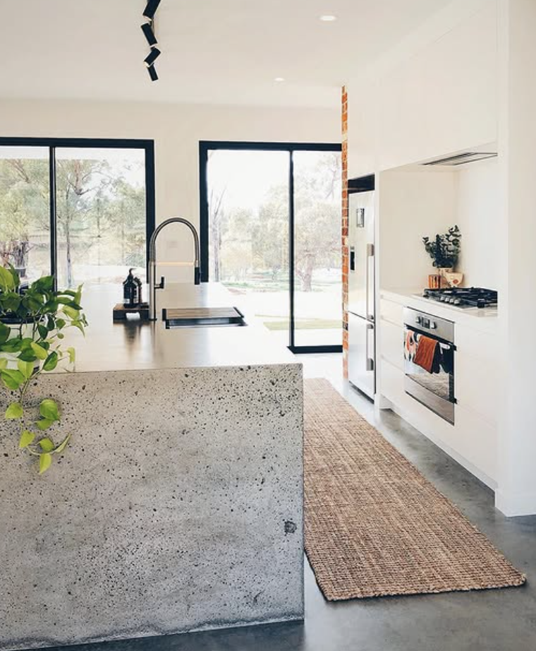 Minimalist kitchen with raw concrete island, sleek black faucet, white cabinetry, and floor-to-ceiling windows.