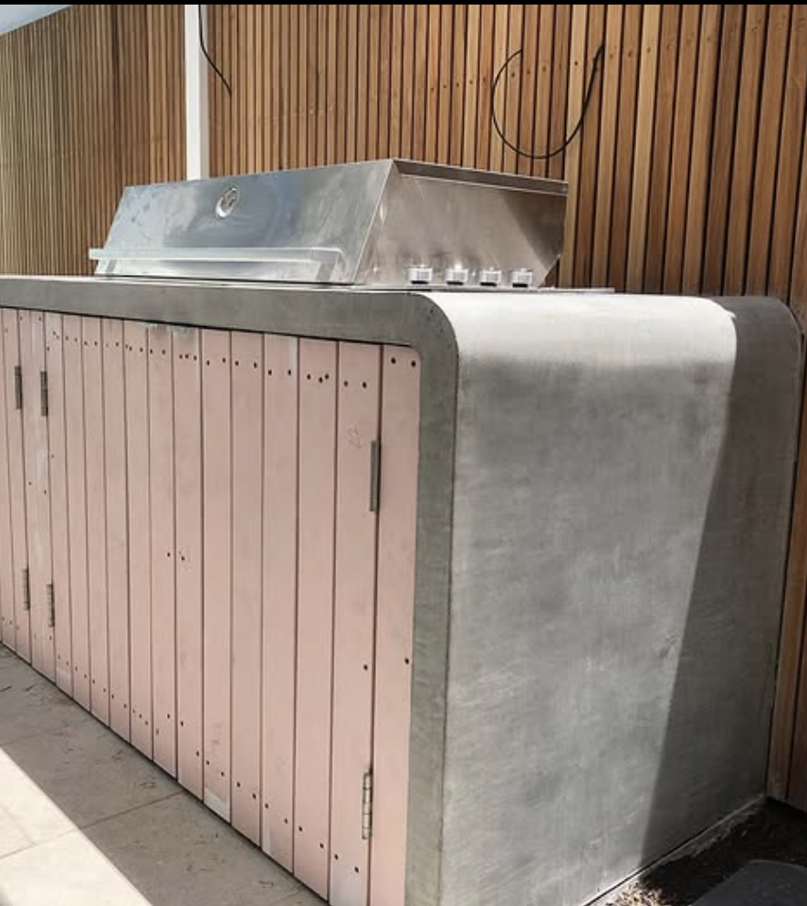 Outdoor kitchen with smooth concrete countertops, curved concrete edges, and pastel pink cabinet doors under a stainless steel grill.