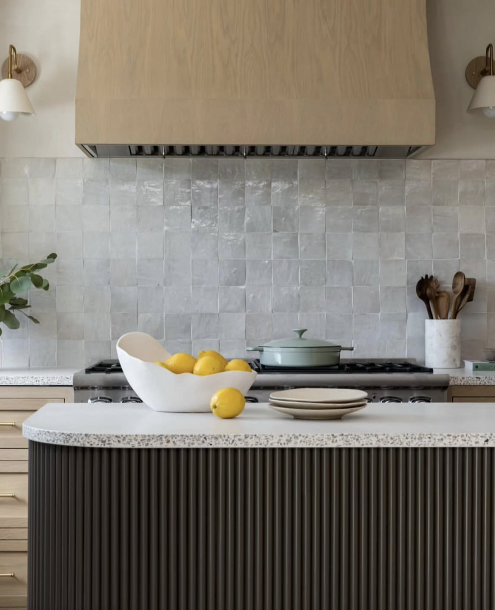 Close-up of a kitchen with a fluted island base, terrazzo-style concrete countertop, light wood range hood, and zellige tile backsplash.