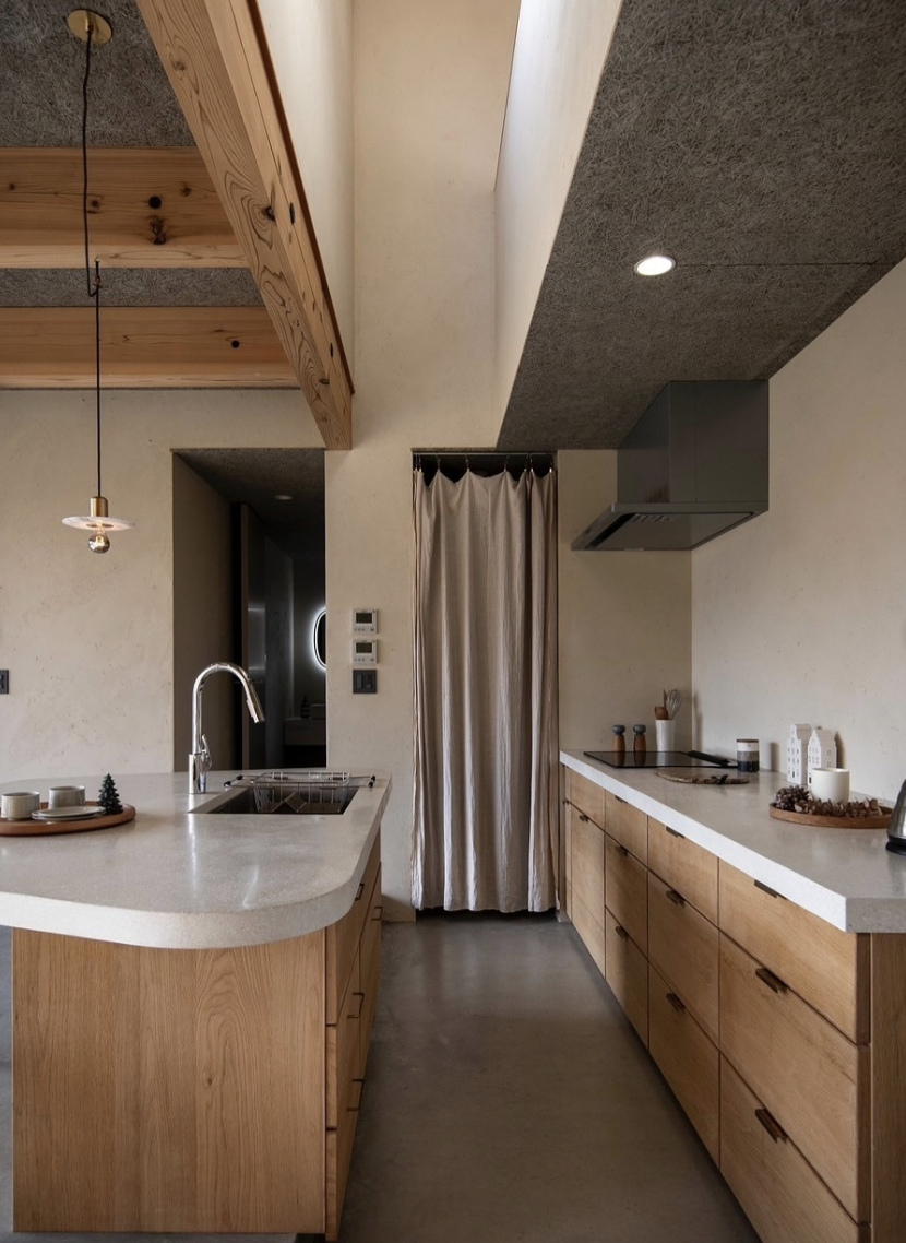 Minimalist kitchen with light wood cabinets, creamy concrete countertops, soft curtain pantry door, and modern pendant lighting.