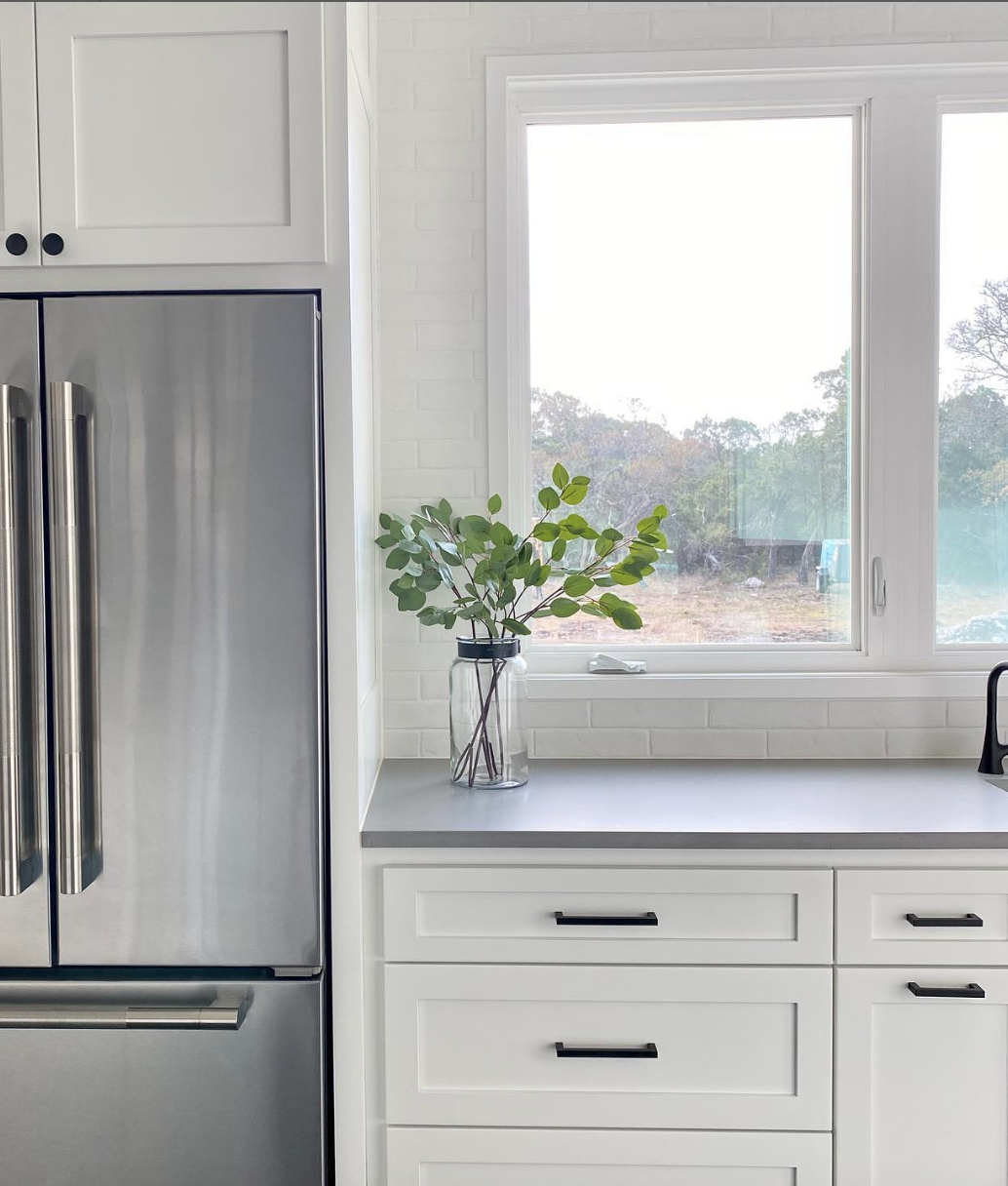 Bright white kitchen with shaker cabinets, gray concrete countertop, black hardware, and stainless steel fridge.