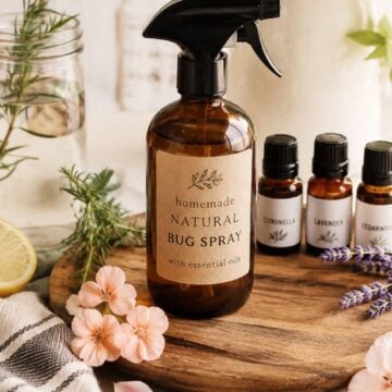 Homemade natural bug spray in an amber glass bottle with citronella, lavender, cedarwood, rosemary, and fresh flowers on a warm farmhouse-style table.