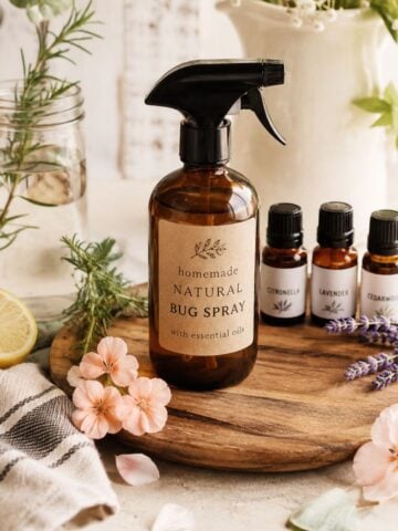 Homemade natural bug spray in an amber glass bottle with citronella, lavender, cedarwood, rosemary, and fresh flowers on a warm farmhouse-style table.