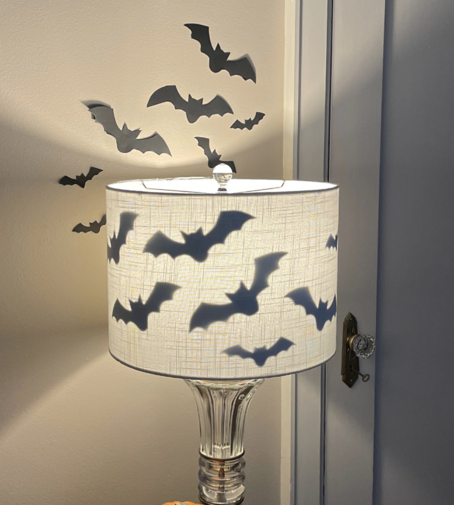Clear glass lamp with white shade casting shadows of black bat cutouts for a spooky DIY Halloween bat decoration.