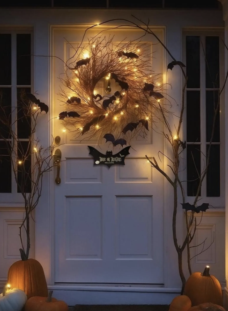 Front door with glowing twig wreath filled with black Halloween bat decorations, pumpkins, and twinkle lights.