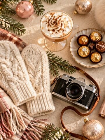 Cozy Christmas desktop wallpaper featuring cream knit mittens, a plaid scarf, vintage camera, hot chocolate, pink ornaments, and evergreen branches.