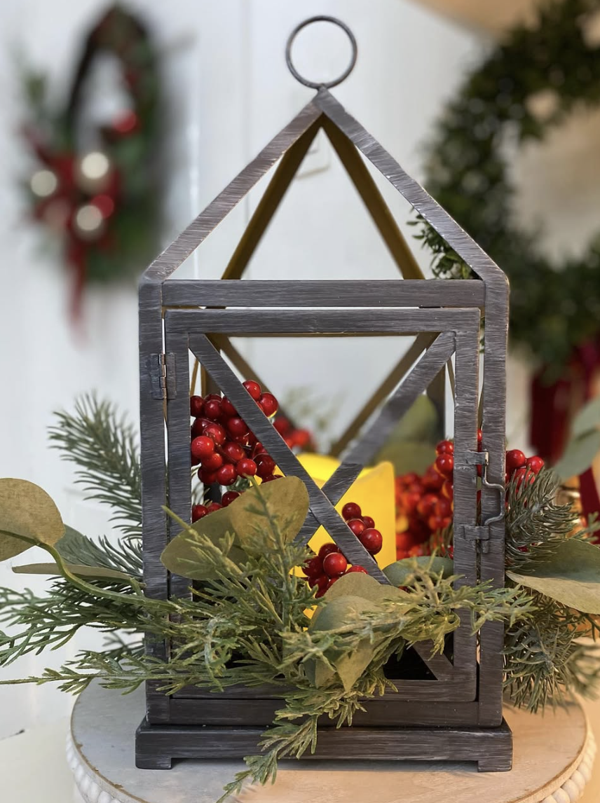 Black farmhouse-style lantern decorated with greenery, red berries, and a glowing candle for Christmas.
